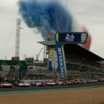 Die Rennwagen stehen am Start der 24 Stunden von Le Mans, während blauer und rosa Rauch in den Himmel aufsteigt und im Hintergrund die Tribünen voller Zuschauer zu sehen sind.